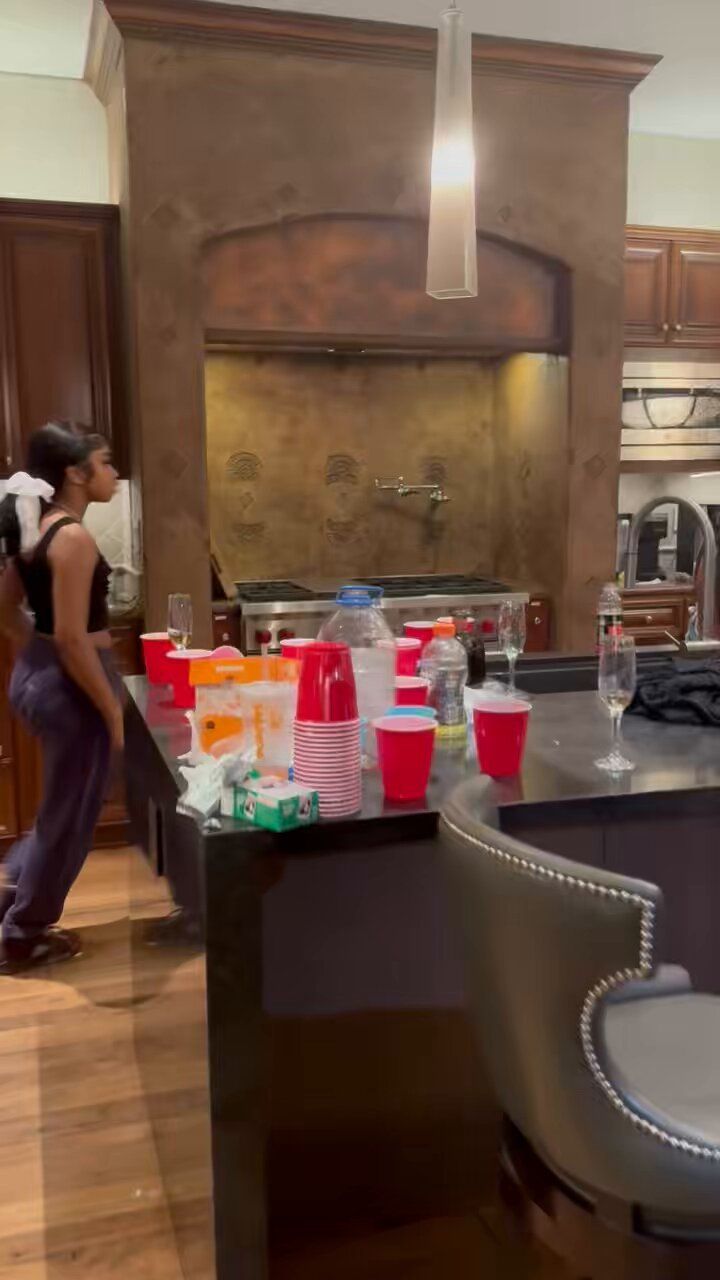 a woman standing in a kitchen next to a counter filled with cups and bottles on it