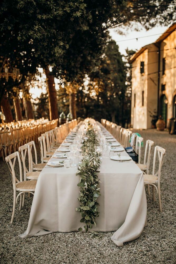 a long table is set with white linens and greenery for an outdoor dinner