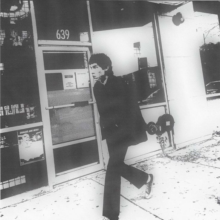 black and white photograph of a man leaning on a gas pump in front of a building