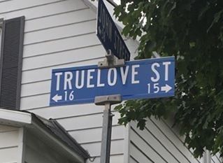 two street signs on the corner of truelov st and 13th ave in front of a house