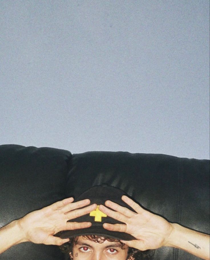 a man sitting on top of a black couch holding his hat over his face with both hands