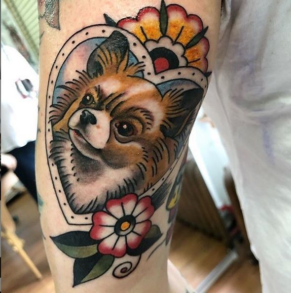 a small dog with flowers on its head in a heart shaped frame tattoo by person