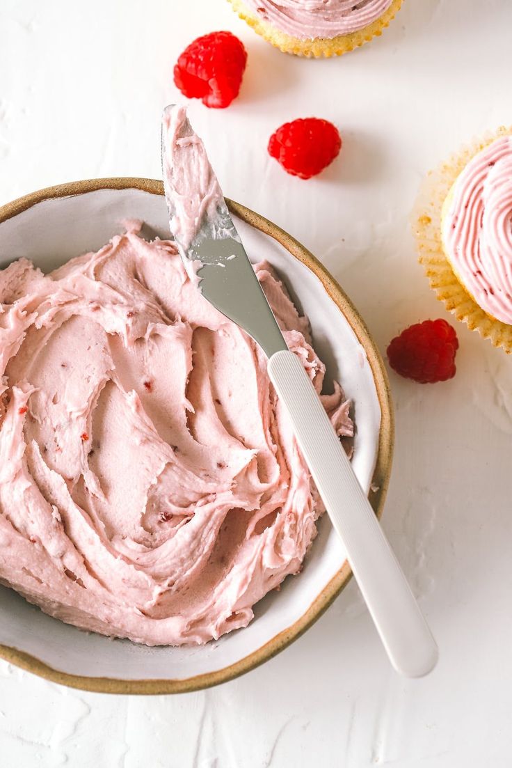 a bowl filled with pink frosting next to cupcakes on a white table