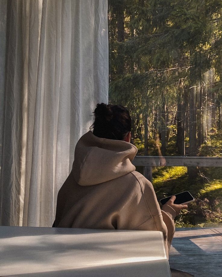 a person sitting in front of a window looking out at the woods while holding a cell phone