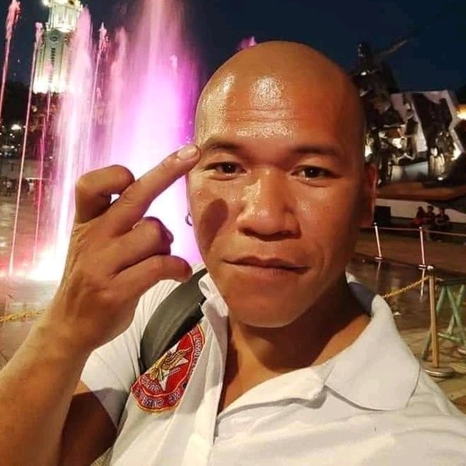 a bald man poses in front of a fountain at night with his finger to his forehead