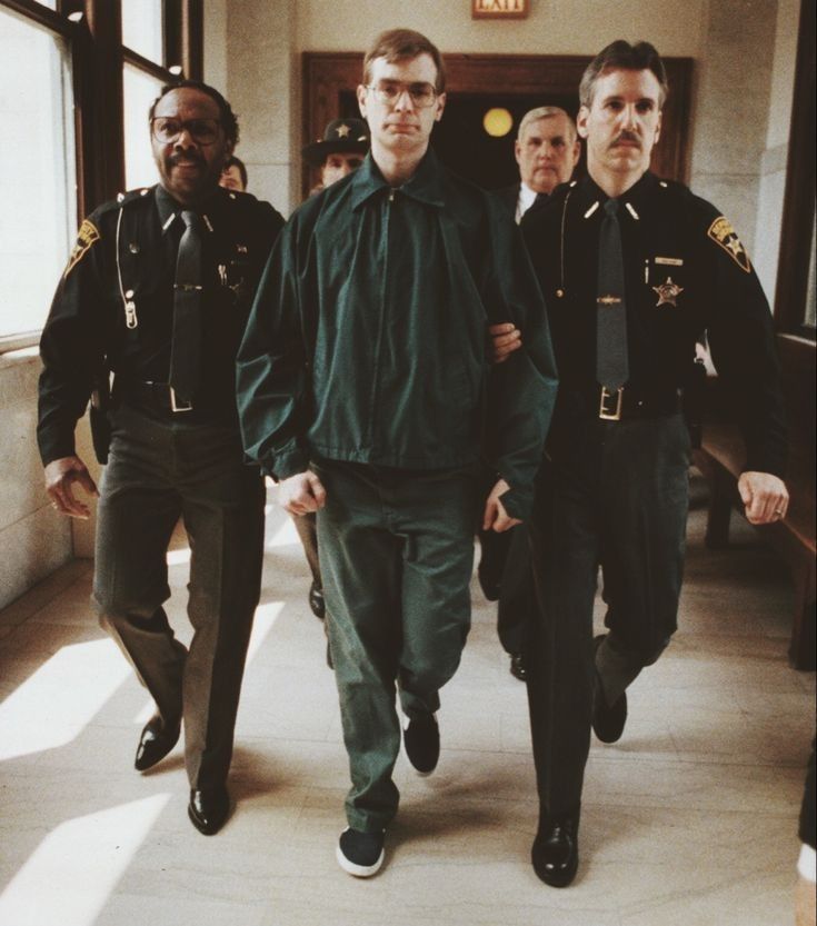 three men in uniform walking down a hall way with one man wearing a green jacket and tie