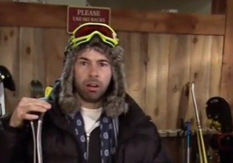 a man wearing a ski hat and goggles in front of a wooden wall with skis on it