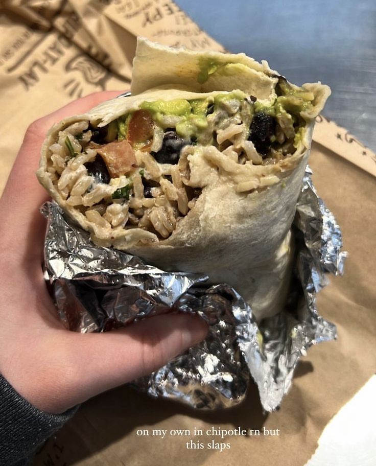 a hand holding a burrito wrapped in aluminum foil on top of a bag filled with food