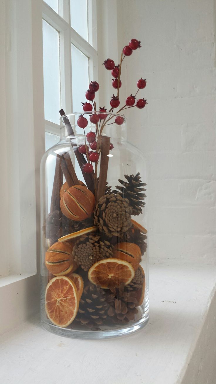 a glass vase filled with oranges and pine cones on top of a window sill