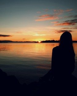 a woman sitting on the shore watching the sun go down