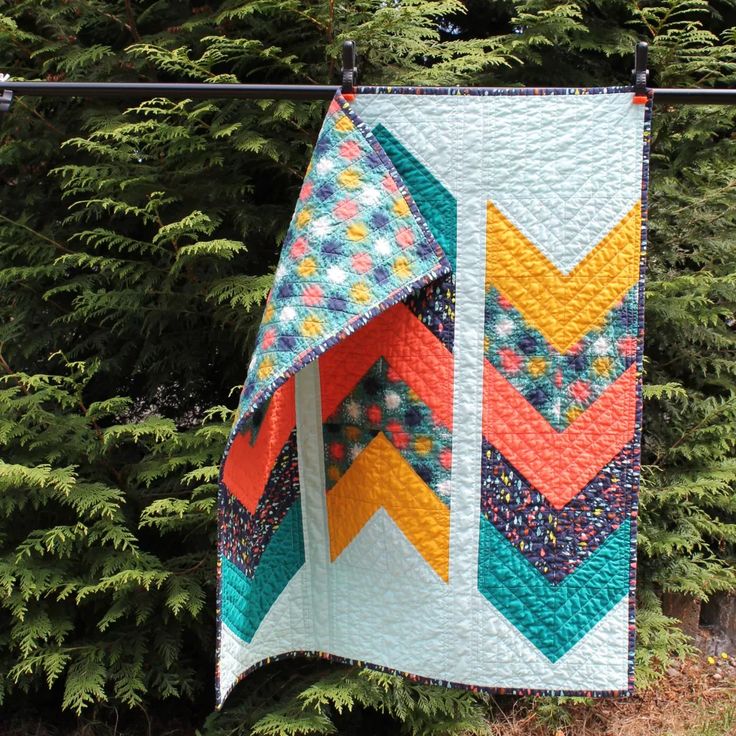 a quilt hanging from a clothes line with trees in the back ground and bushes behind it