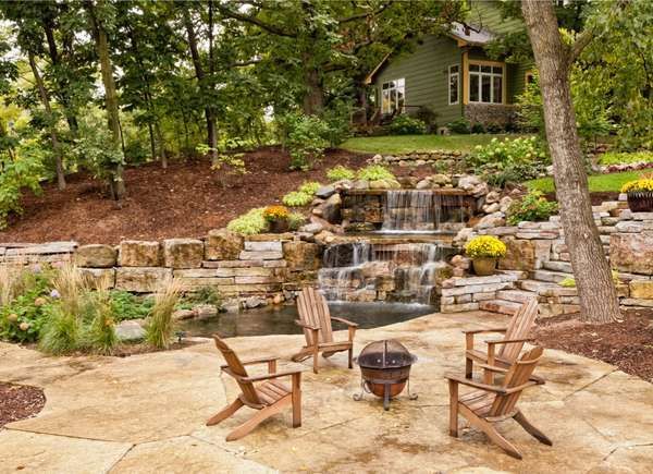 a patio with chairs and a fire pit in front of a waterfall surrounded by trees