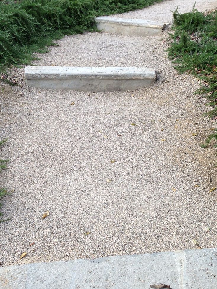 a cement bench sitting on top of a sidewalk next to green grass and bushes in front of it