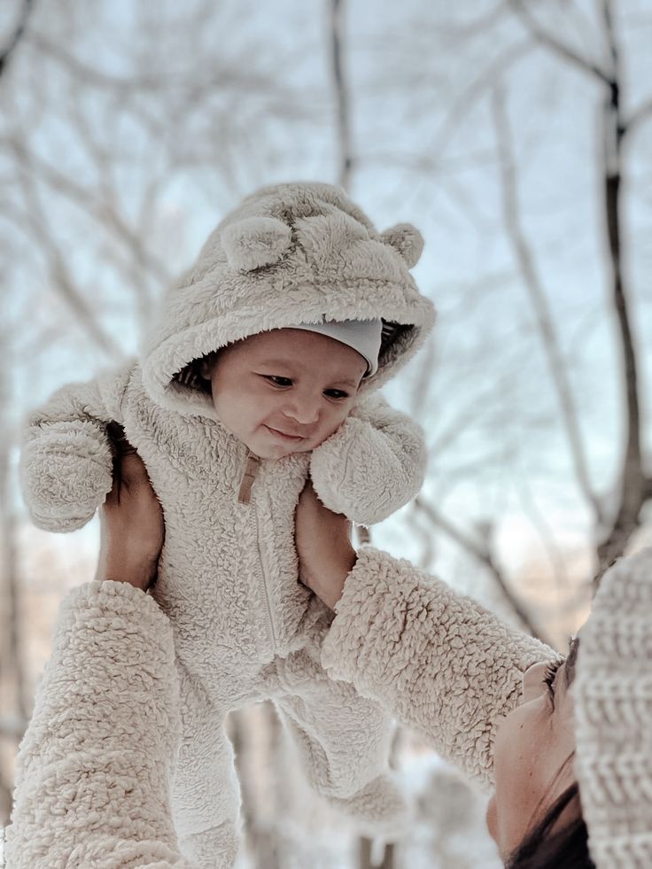 a woman holding a baby in the snow wearing a teddy bear suit and mittens