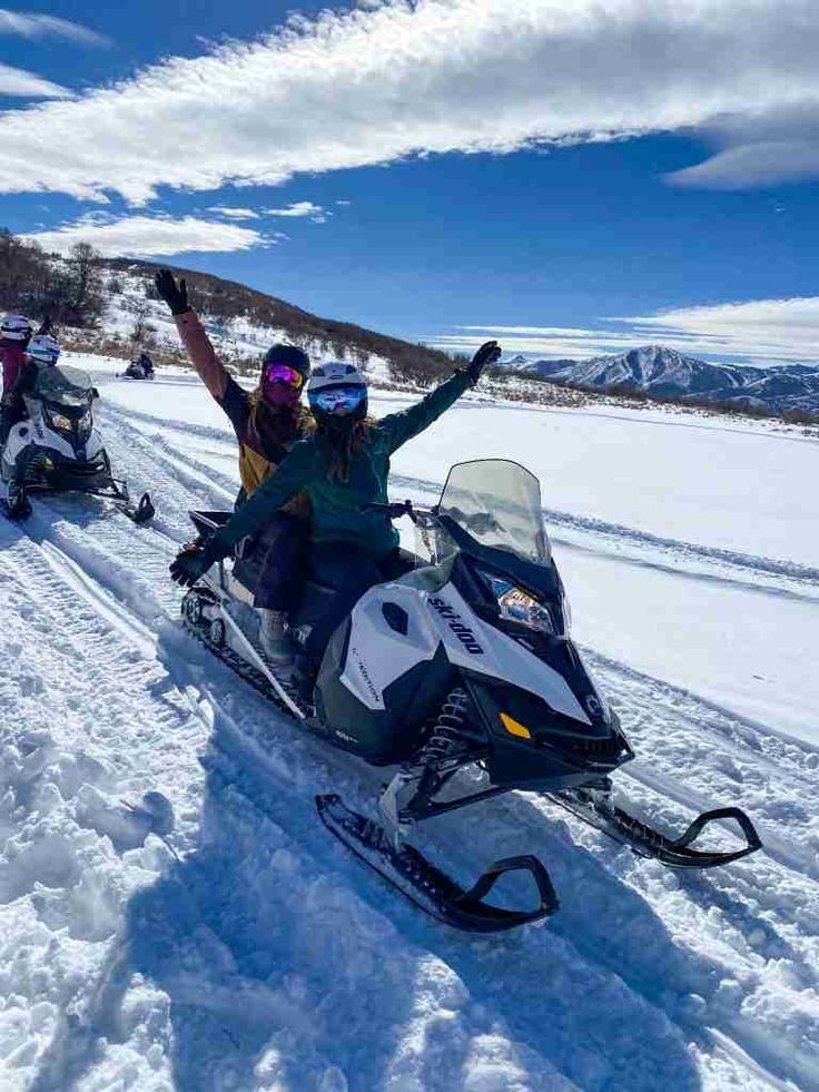 three people riding snowmobiles in the snow with their arms up and one person on skis