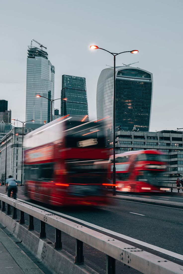 two red double decker buses driving down a street next to tall buildings in the city