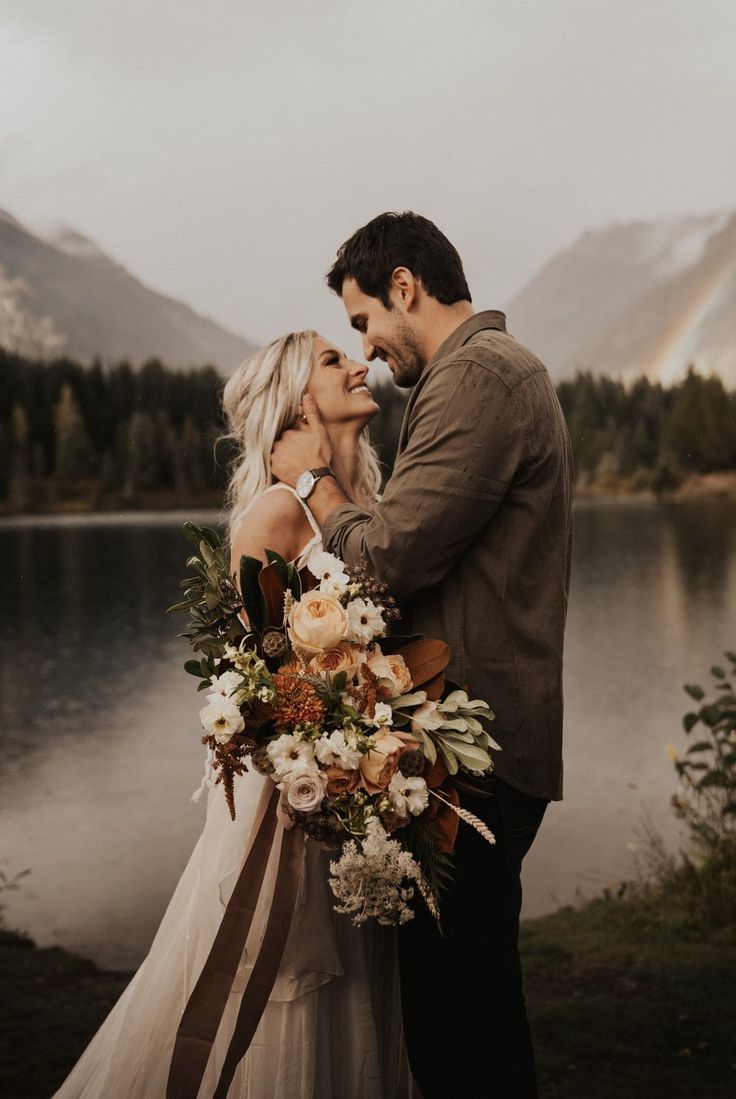 a couple standing next to each other in front of a lake with mountains and trees