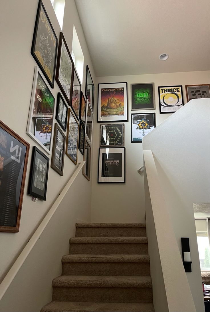 the stairs are lined with framed pictures and posters on the wall above them is a staircase leading up to an upstairs landing area