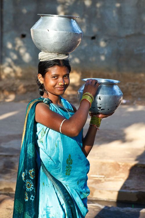 a woman carrying a pot on her head and holding a silver bowl on her shoulder