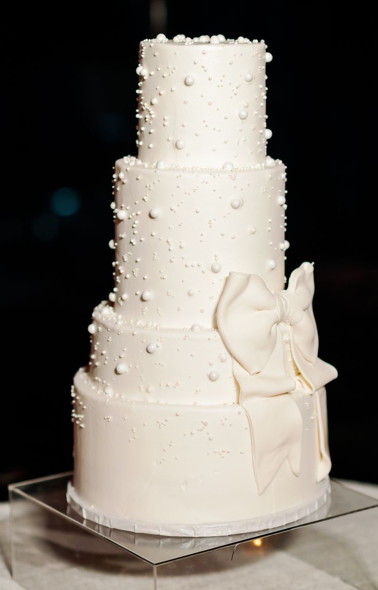 a three tiered white wedding cake with pearls and bows on the top, sitting on a clear acrylic stand