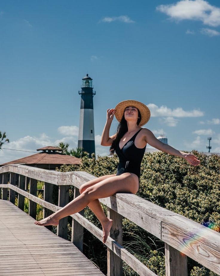 a woman in a black swimsuit and straw hat sitting on a wooden bridge near a light house