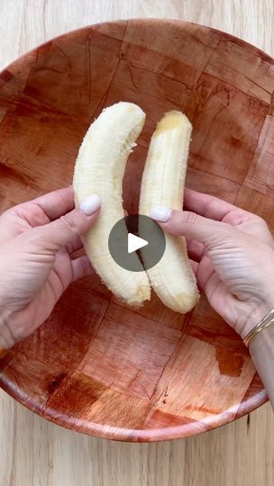 two hands holding bananas in a wooden bowl on top of a wood table, with the banana split open