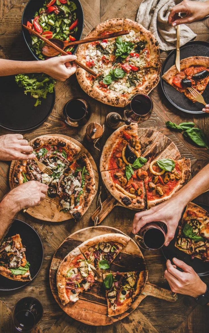 several people are eating pizza and drinking wine at a table with many different types of food