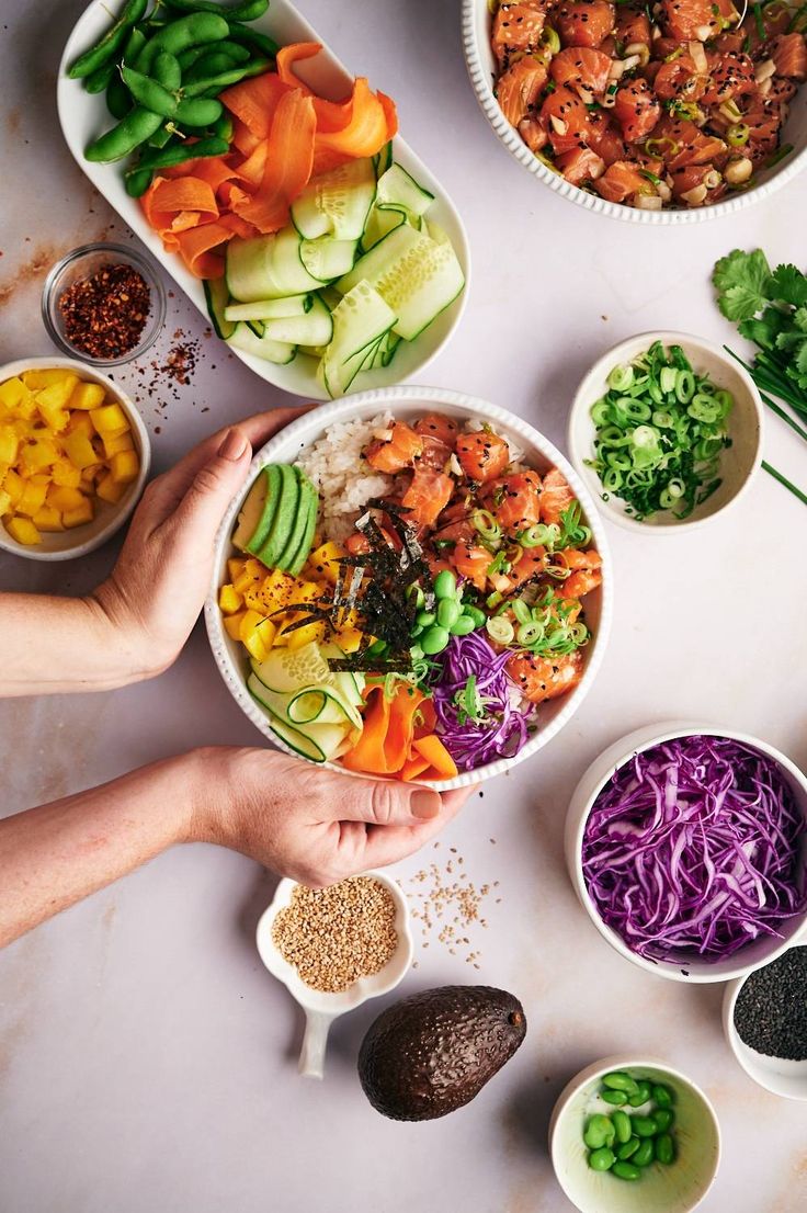 hands holding bowls filled with food on top of a white countertop next to bowls of vegetables and other foods