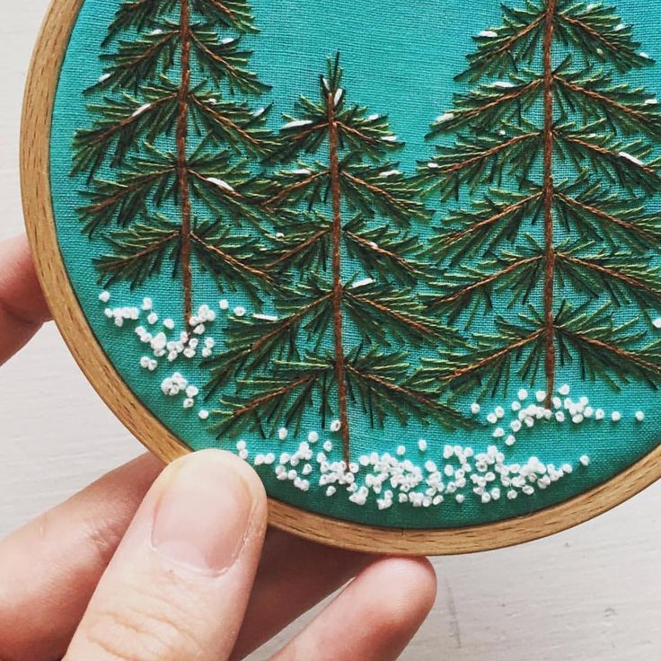 someone is holding up a small embroidery project with pine trees in the snow on it