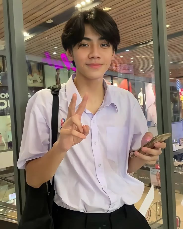 a young man in a white shirt is holding a cell phone and giving the peace sign