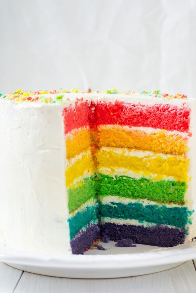 a rainbow cake on a white plate with one slice cut out