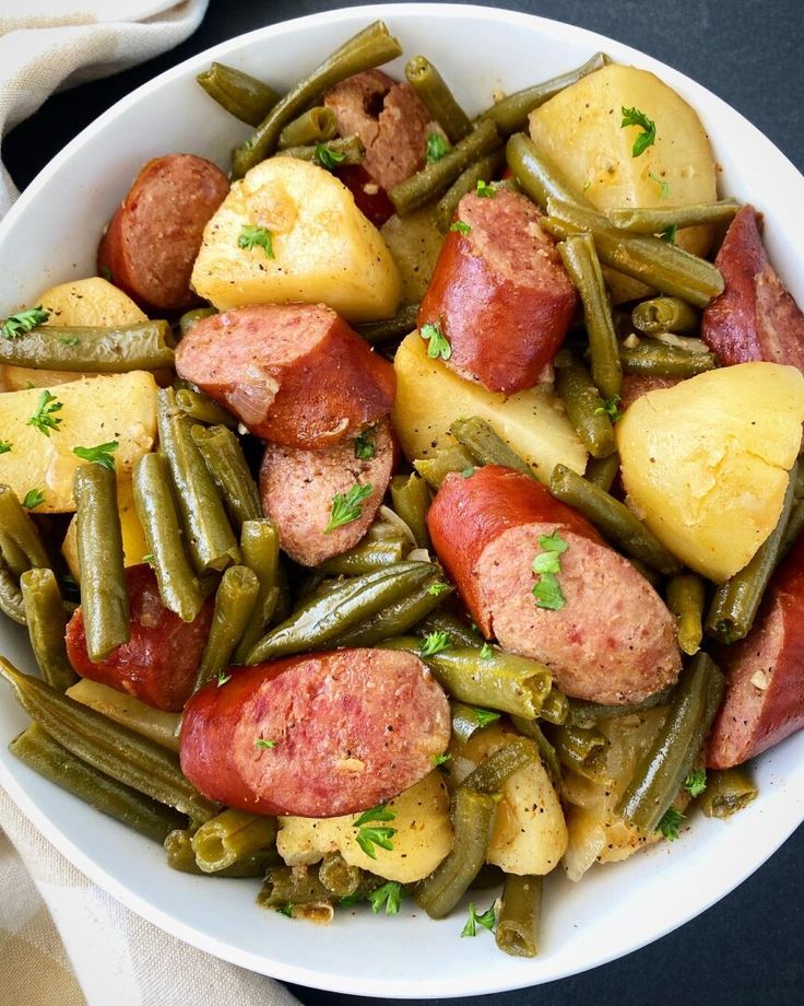 a white bowl filled with green beans, potatoes and kielbasa on top of a black table