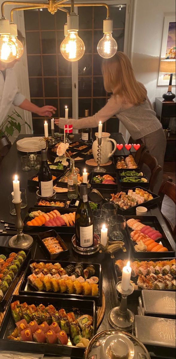 two people standing at a table with many trays of food on it and candles in front of them