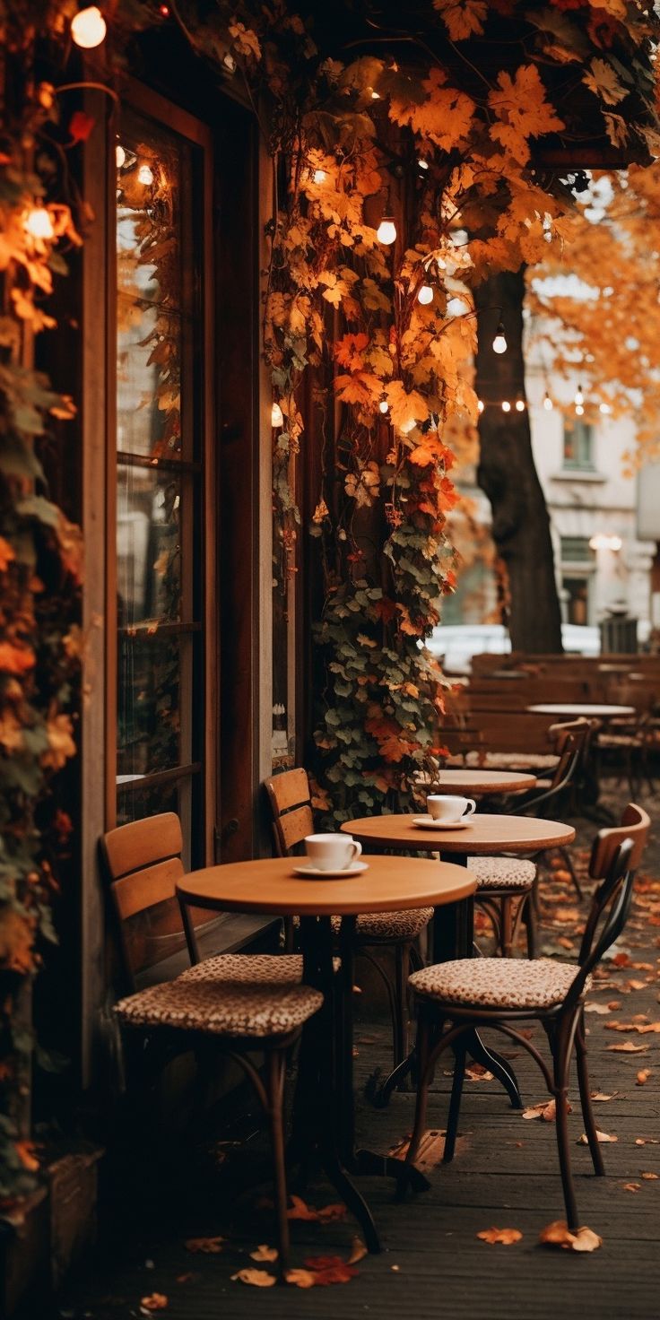 an outdoor cafe with tables and chairs covered in autumn leaves, next to a tree