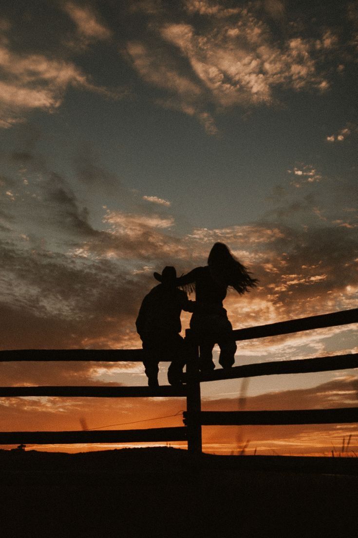 two people sitting on top of a wooden fence under a cloudy sky with the sun setting behind them