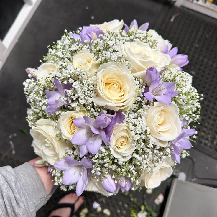 a bouquet of white and purple flowers is being held by someone's hand on the ground