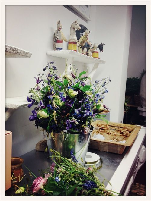 purple flowers are in a silver bucket on a kitchen counter next to a cutting board