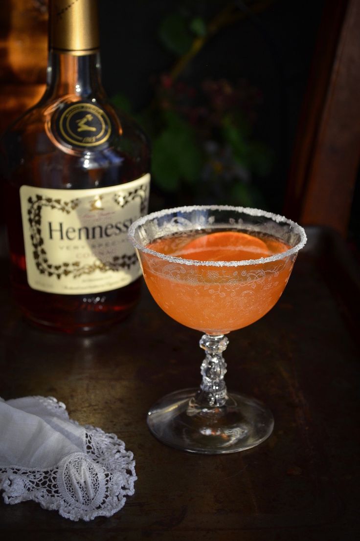 an orange cocktail in a coupe glass next to a bottle of hennesson's