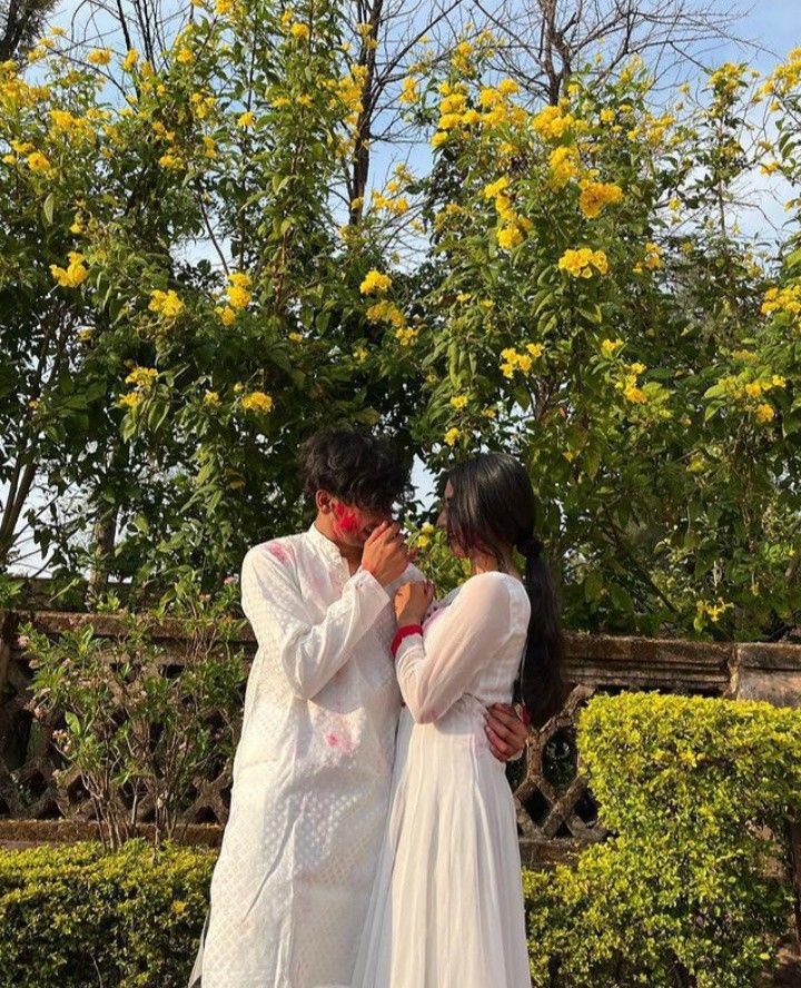 two people standing next to each other in front of bushes and trees with yellow flowers