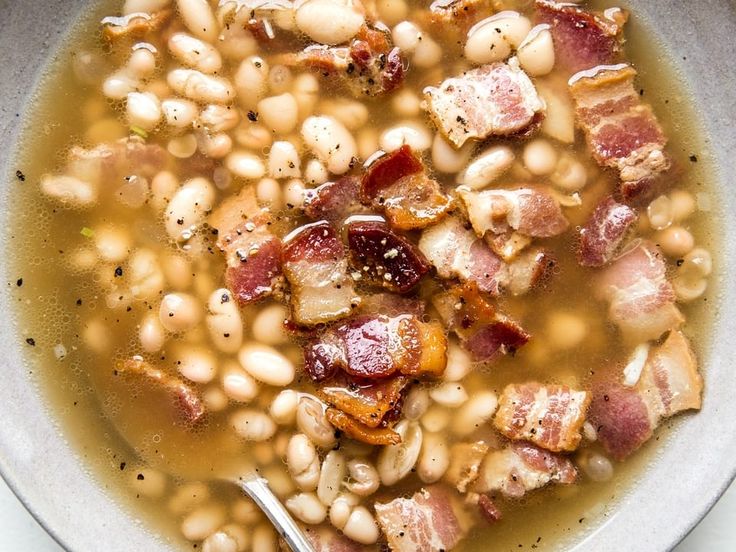 a white bowl filled with beans and bacon next to a slice of toasted bread