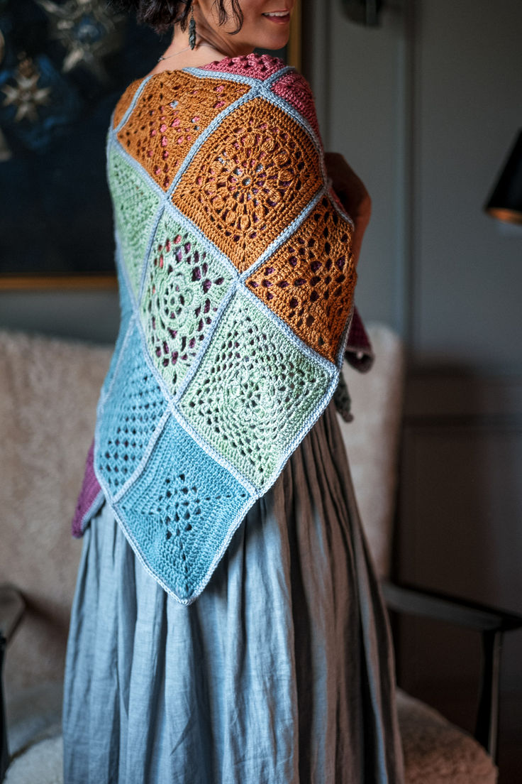 a woman wearing a colorful crocheted shawl in front of a couch with a painting on the wall behind her