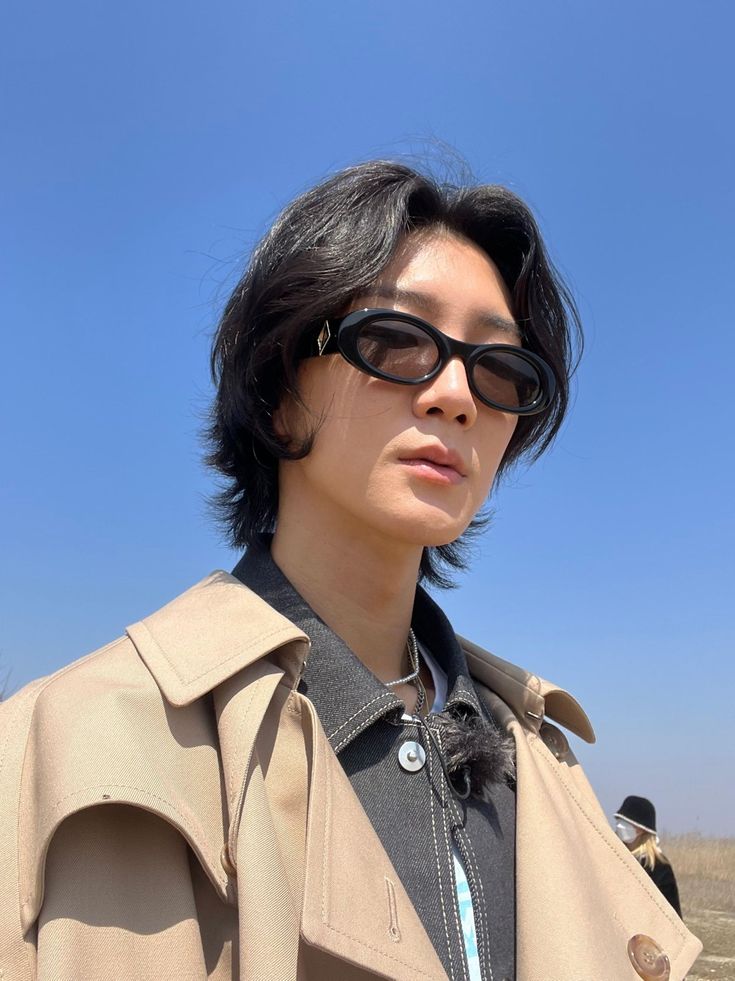 a woman wearing sunglasses and a trench coat looks off into the distance while standing in an open field