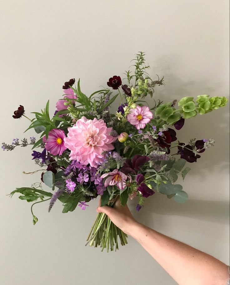 a hand holding a bouquet of flowers against a white wall with purple and green foliage