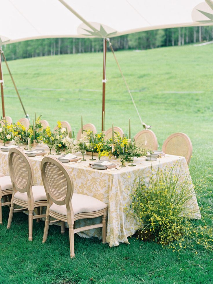 an outdoor table set up with chairs and tables covered in yellow cloths, surrounded by greenery