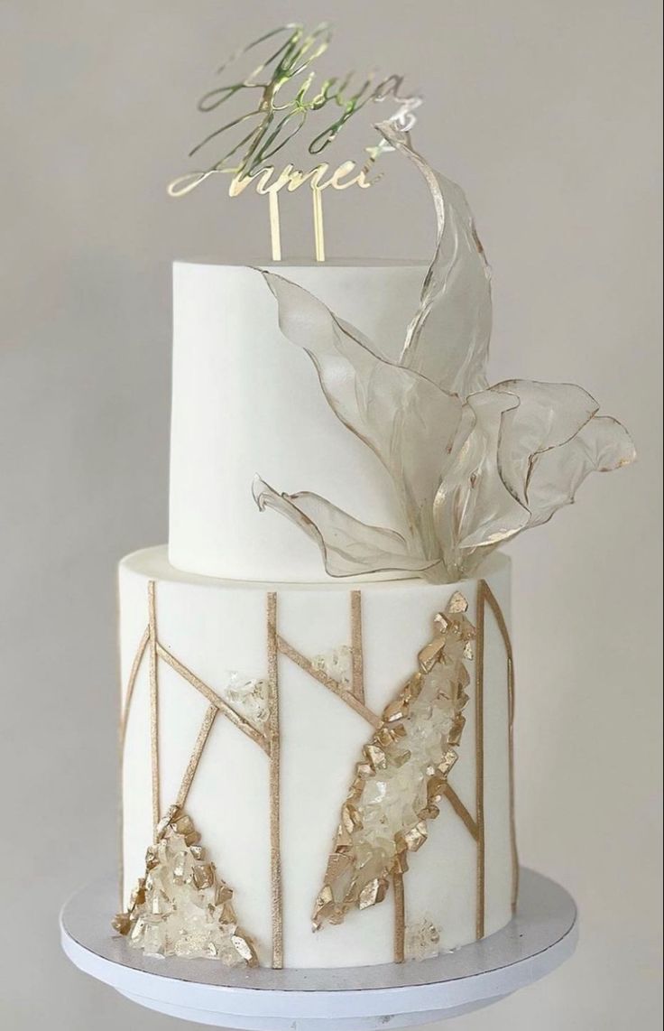 a white and gold wedding cake with flowers on the top is topped with an acrylic leaf