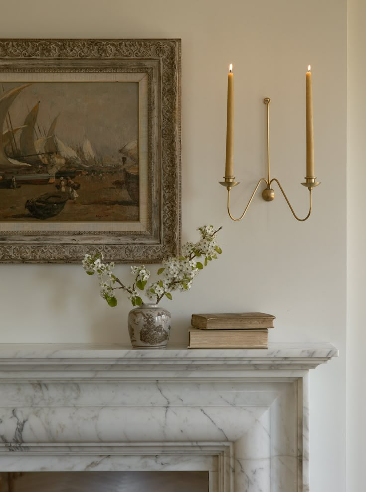 a white fireplace with two candles and a painting on the wall above it that has flowers in a vase