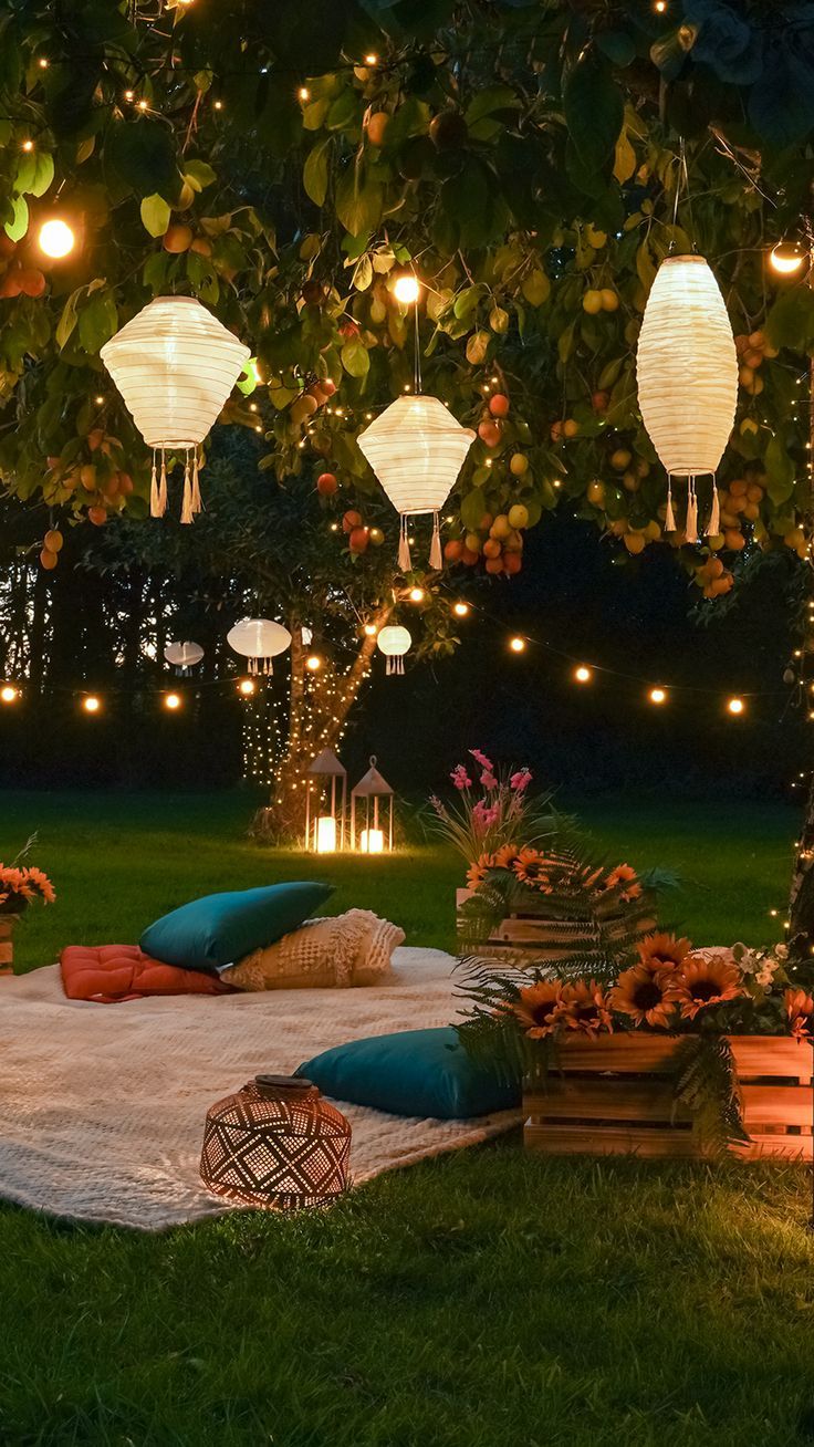 an outdoor seating area with lanterns and pillows on the grass, surrounded by flowers and trees