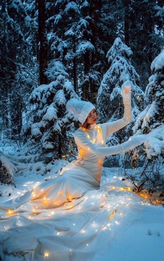 a woman dressed in white sitting on the ground surrounded by snow covered trees and lights