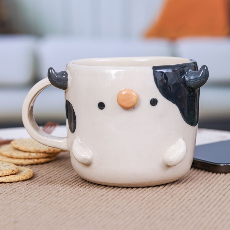 a cow mug sitting on top of a table next to crackers and a cell phone