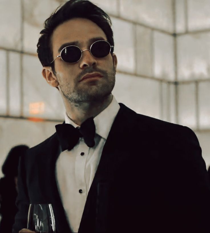 a man in a tuxedo holding a wine glass and looking at the camera